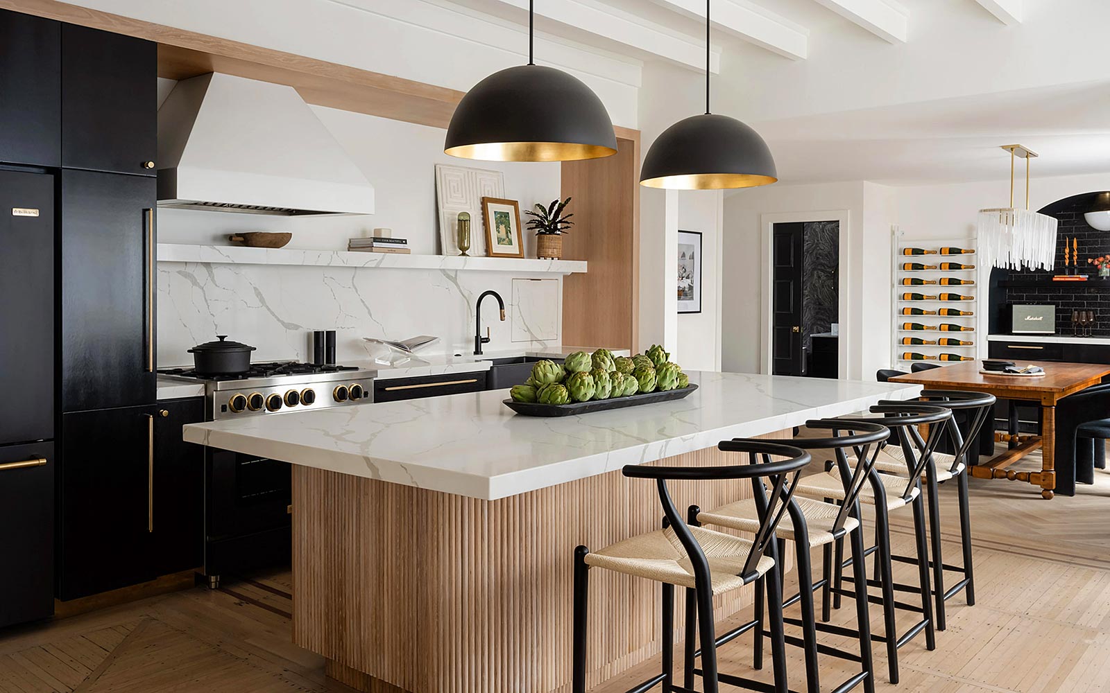 A wide view into the kitchen across a large island countertop to the stove and sink on the far wall. Two large pendant lamps hang painted with black exterior and reflective gold interior above the white marble island.